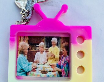 May include: A pink and yellow television-shaped keychain with a picture of four women inside. The keychain has a silver charm attached to it.