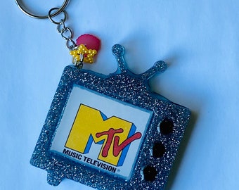 May include: A silver glitter television keychain with a blue and yellow MTV logo and the text "MUSIC TELEVISION". The keychain has a silver key ring and a pink and yellow beaded charm.