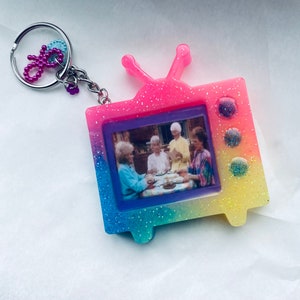 May include: A colorful, glittery TV-shaped keychain with a picture of four women sitting at a table. The keychain is pink, blue, yellow, and green.