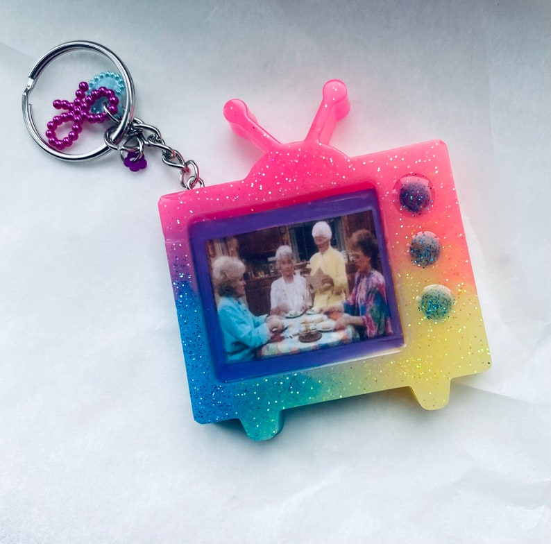 May include: A colorful, glittery TV-shaped keychain with a picture of four women sitting at a table. The keychain is pink, blue, yellow, and green.