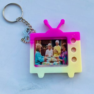 May include: A pink and yellow television-shaped keychain with a picture of four women inside. The keychain has a silver key ring with a small charm attached.