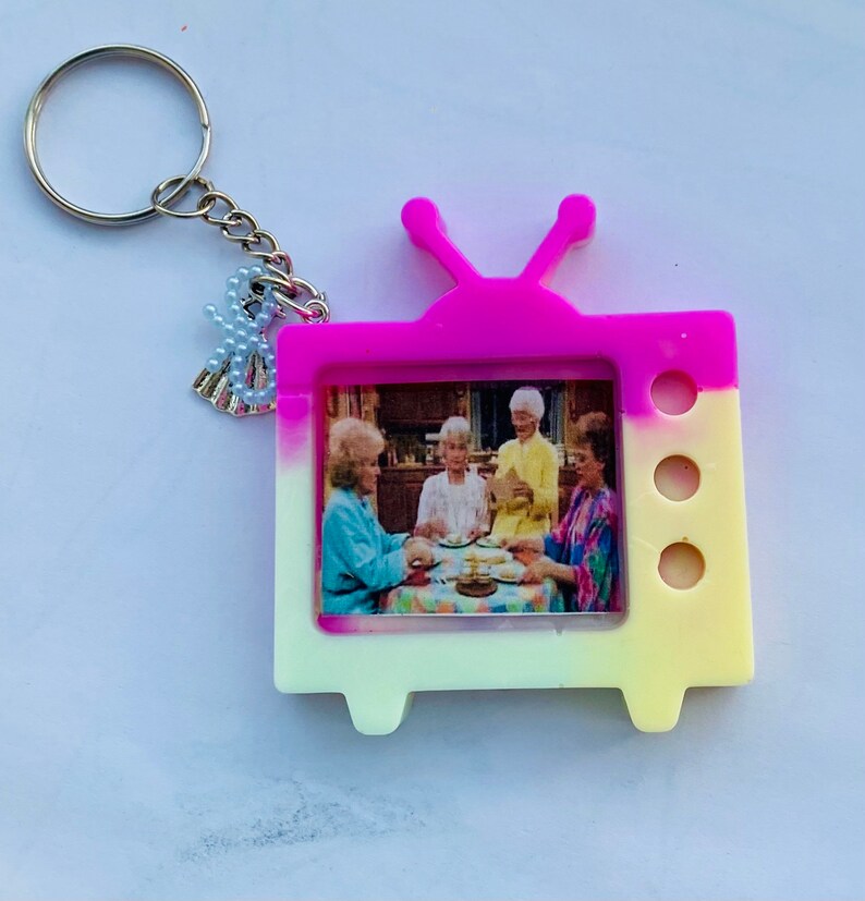 May include: A pink and yellow television-shaped keychain with a picture of four women inside. The keychain has a silver key ring with a small charm attached.