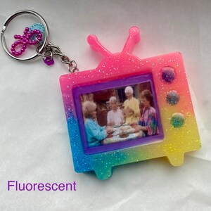 May include: A pink, yellow, and blue glittery television keychain with a picture of four women sitting at a table. The keychain has a silver key ring attached.