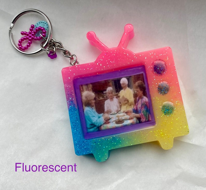 May include: A pink, yellow, and blue glittery television keychain with a picture of four women sitting at a table. The keychain has a silver key ring attached.