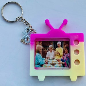 May include: A pink and yellow television-shaped keychain with a picture of four women inside. The keychain has a silver charm attached to it.