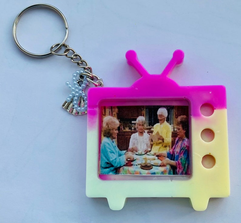 May include: A pink and yellow television-shaped keychain with a picture of four women inside. The keychain has a silver charm attached to it.