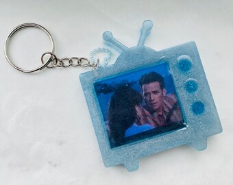 May include: A light blue glitter keychain shaped like a vintage television with a picture of a couple inside. The television has three blue glitter dots on the side.