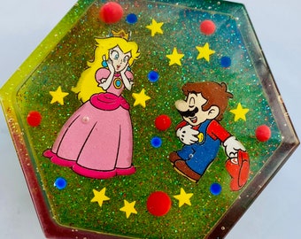 May include: A hexagonal resin trinket dish with a glittery green background. The dish features a cartoon image of Princess Peach and Mario from the Super Mario Bros. video game series. The image is surrounded by yellow stars and red and blue dots.
