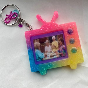 May include: A pink, blue, and yellow glittery TV-shaped keychain with a picture of four women inside. The keychain has a silver chain and a pink beaded charm.