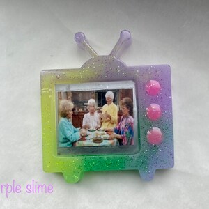 May include: A glittery purple, green, and yellow TV-shaped resin pin with a picture of four women inside. The pin has three pink circles on the right side.