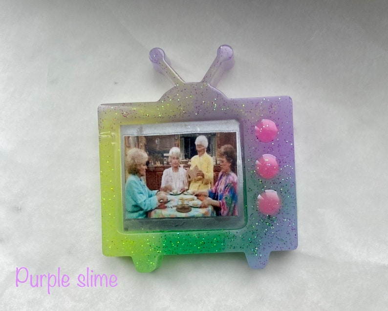 May include: A glittery purple, green, and yellow TV-shaped resin pin with a picture of four women inside. The pin has three pink circles on the right side.