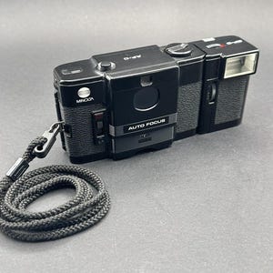 Könnte beinhalten: Schwarze Minolta AF-D Kamera mit eingebautem Blitz und schwarzem geflochtenem Handgelenkgurt. Auf der Kamera stehen die Wörter "Minolta" und "Auto Focus". Die Kamera liegt auf einer grauen Oberfläche.