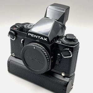 Puede incluir: Una cámara Pentax negra con un accesorio de flash. La cámara tiene una tapa de lente y un agarre texturizado. La palabra "PENTAX" está impresa en blanco en el cuerpo de la cámara. La cámara está sobre un fondo blanco.