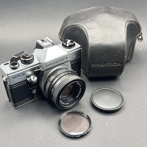 May include: A vintage Praktica MTL 5 camera with a Pentacon lens, lens cap, and filter. The camera is black and silver, with the brand name visible. A black leather case is also present.