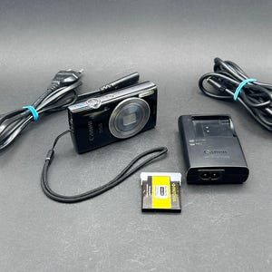 Op de afbeelding: Een zwarte Canon IXUS digitale camera met polsband, een zwarte Canon acculader, een geel met zwarte Patona oplaadbare accu en twee zwarte stroomkabels met blauwe tie-wraps, allemaal gerangschikt op een donkergrijs oppervlak.