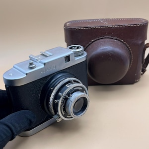 Puede incluir: Una cámara Ajax D vintage en negro y plata con una funda de cuero marrón. La cámara tiene una parte superior y detalles de la lente plateados, con el nombre "Ajax D" visible. La lente tiene anillos ajustables. La funda de cuero marrón está en el fondo.