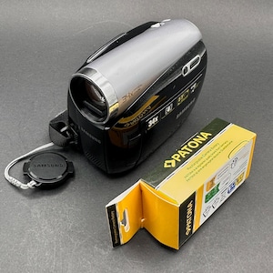 May include: A black and silver Samsung camcorder with a 34x optical zoom lens. The lens cap is attached with a strap. A yellow and black Patona rechargeable camera battery box is open next to the camcorder.