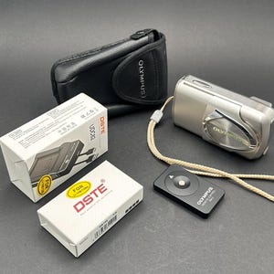 May include: A silver Olympus digital camera with a black leather case, a remote control, and two white boxes branded DSTE. The camera has a beige strap. The boxes have the text "FOR Li-15B/12B".