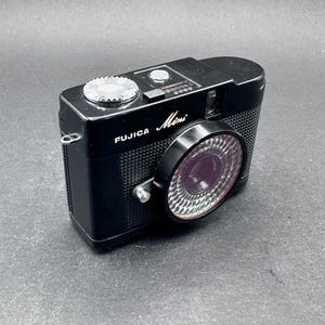 以下が含まれることがあります： 黒いFujica Miniフィルムカメラ。テクスチャ加工されたボディ、銀色の模様入りリングが付いた円形のレンズハウジング、上部の銀色のダイヤル、小さな長方形のファインダー窓が特徴です。ブランド名とモデル名が白文字で印字されています。