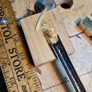 May include: A woodworking scene featuring a wooden block with a gouge tool carving out wood shavings. A vintage ruler with the text "BRISTOL STORE INC" is visible. The gouge tool is black and the wood is light brown.
