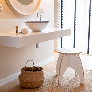 Può includere: Un mobile da bagno bianco con un lavabo in ceramica bianca e un rubinetto cromato. Un cesto intrecciato si trova sul pavimento sotto il mobile. Uno sgabello bianco con un design a croce si trova su un tappeto intrecciato di fronte al mobile.