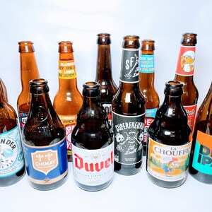 May include: A collection of 11 brown glass beer bottles with various labels. The labels feature different colours, designs, and text, including "Lowlander White Ale", "Chimay", "Duvel", "Superfreunde", "La Chouffe", and "Del IPA".