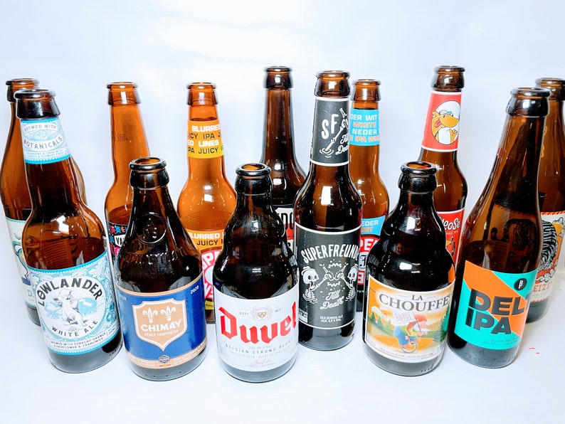 May include: A collection of 11 brown glass beer bottles with various labels. The labels feature different colours, designs, and text, including "Lowlander White Ale", "Chimay", "Duvel", "Superfreunde", "La Chouffe", and "Del IPA".