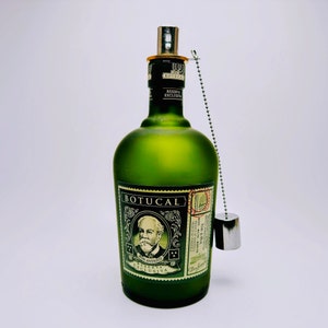 May include: A frosted green glass bottle of Botucal Reserva Exclusiva rum featuring a portrait of a man with a mustache on the label. The bottle has a silver metal pourer attached to the neck by a silver ball chain with a small silver cap hanging.