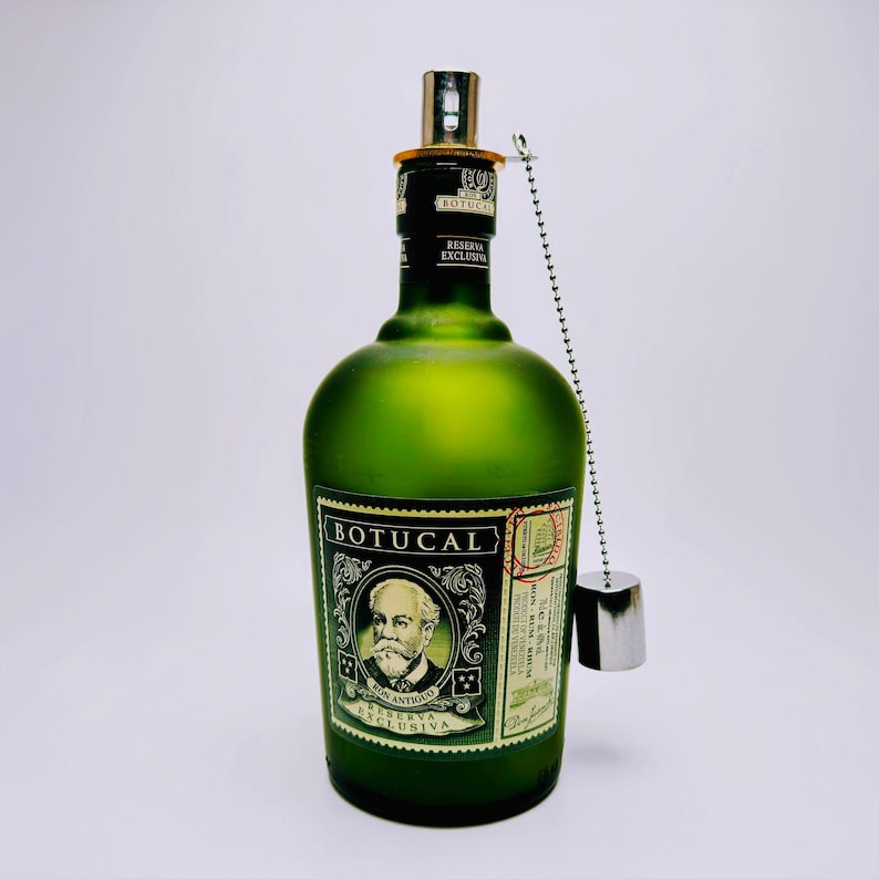 May include: A frosted green glass bottle of Botucal Reserva Exclusiva rum featuring a portrait of a man with a mustache on the label. The bottle has a silver metal pourer attached to the neck by a silver ball chain with a small silver cap hanging.
