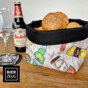 Könnte beinhalten: Ein schwarzer und weißer Stoff-Brotkorb mit schwarzem Besatz. Der Korb ist mit Brötchen gefüllt und hat einen Aufdruck von Bierflaschen und dem Text "BIER ZEUG".