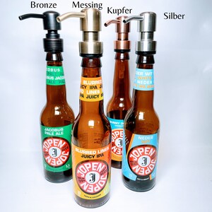 May include: Four glass bottles with different coloured labels and pump tops. The labels have text in Dutch and English. The pump tops are bronze, brass, copper, and silver.