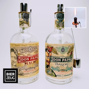 May include: Two clear glass bottles with labels that say "Don Papa Rum" and "Don Papa Baroko". The bottles have a silver cap and a silver chain with a silver charm.