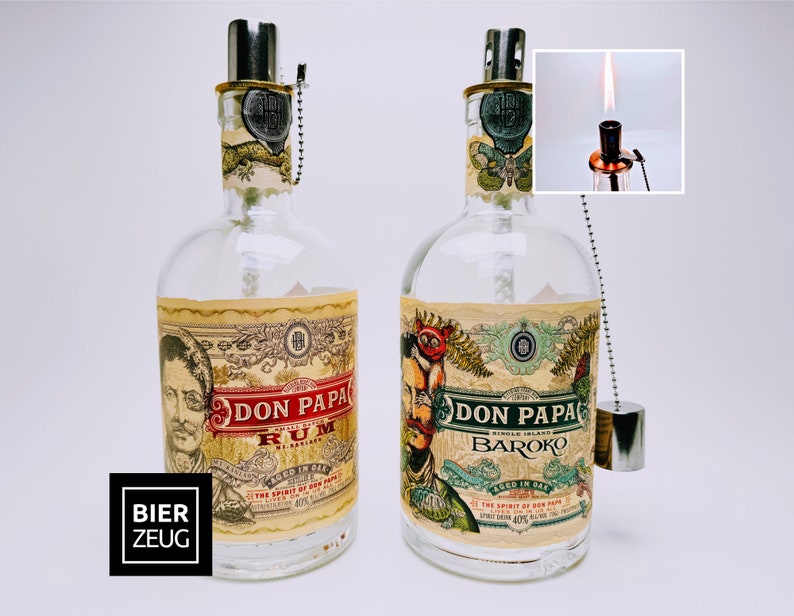 May include: Two clear glass bottles with labels that say "Don Papa Rum" and "Don Papa Baroko". The bottles have a silver cap and a silver chain with a silver charm.