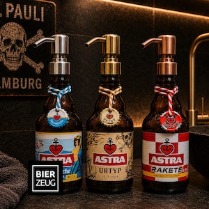 Könnte beinhalten: Drei Seifenspender aus braunen Bierflaschen mit Pumpaufsätzen in Silber, Gold und Kupfer. Jede Flasche hat ein farbiges Etikett mit dem Astra-Logo, einem Herz-Anker-Symbol und einem dekorativen Kronkorken-Anhänger an einer Kordel.
