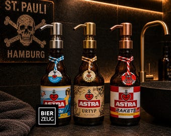 May include: Three glass soap dispensers made from brown beer bottles with pump tops in silver, gold, and copper finishes. Each bottle features a colorful label with the Astra brand logo, a heart-anchor icon, and a decorative bottle cap tag.