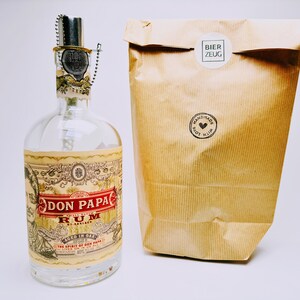 May include: A clear glass bottle with a label that reads "Don Papa Rum" and "Aged in Oak". The bottle is sitting on a white surface next to a brown paper bag with a white sticker that reads "Bier Zeug" and a black sticker that reads "Handmade with Love".