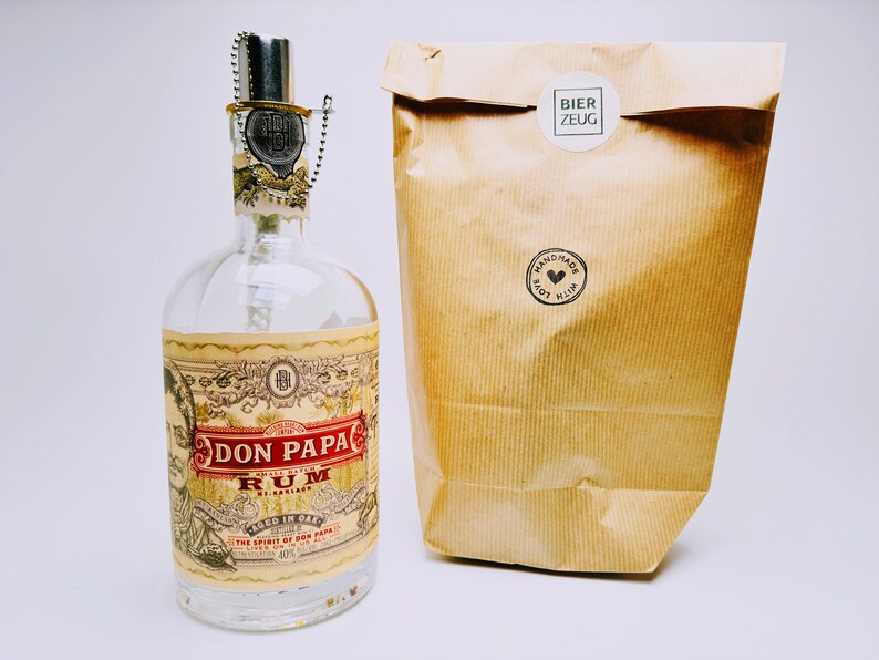 May include: A clear glass bottle with a label that reads "Don Papa Rum" and "Aged in Oak". The bottle is sitting on a white surface next to a brown paper bag with a white sticker that reads "Bier Zeug" and a black sticker that reads "Handmade with Love".