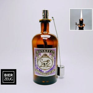 Könnte beinhalten: Eine braune Glasflasche mit einem violetten Etikett, auf dem "MONKEY 47 Schwarzwald Dry Gin" steht. Das Etikett zeigt eine Affenillustration und eine Krone. Die Flasche hat eine silberne Kette, die an der Oberseite befestigt ist, und einen silbernen Flaschenverschluss.