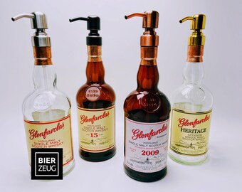 May include: Four decorative liquor bottles with pump dispensers. The bottles are labeled "Glenfarclas" and "Heritage" with various years and styles. The dispensers have silver, black, copper, and gold finishes. The bottles are on a white background.