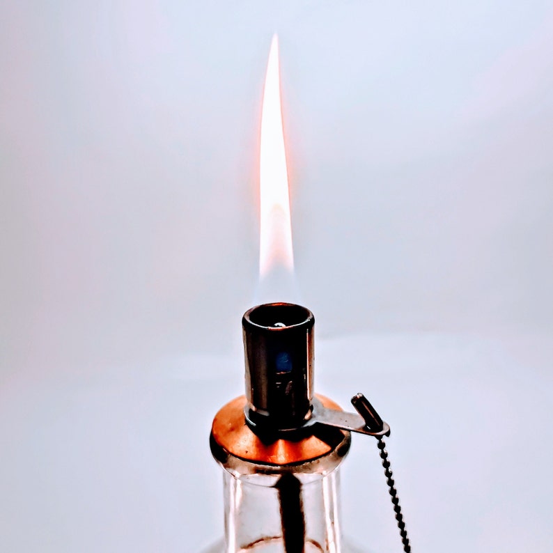 May include: A clear glass bottle with a metal top and a flame burning from the top. The bottle has a copper colored base.