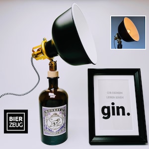 Könnte beinhalten: Eine schwarze und goldene Lampe mit einem schwarzen Schirm ist auf einer braunen Glasflasche mit einem Etikett mit der Aufschrift "Monkey 47" montiert. Die Lampe ist an ein schwarz-weißes Kabel angeschlossen. Ein gerahmter Druck mit dem Text "Gib deinem Leben einen gin." befindet sich auf einer weißen Oberfläche.