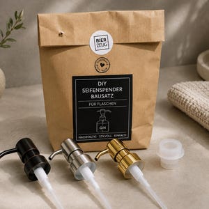 Può includere: Un sacchetto di carta marrone etichettato DIY SEIFENSPENDER BAUSATZ FÜR FLASCHEN con tre erogatori di sapone in metallo con finiture nero opaco, argento e oro, più un adattatore in plastica bianca, disposti davanti al sacchetto su una superficie.