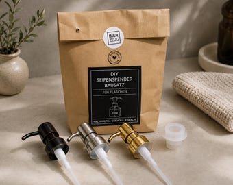 May include: A brown paper bag labeled DIY SEIFENSPENDER BAUSATZ FÜR FLASCHEN with three metal soap pump dispensers in matte black, silver, and gold finishes, plus a white plastic adapter, arranged in front of the bag on a flat surface.