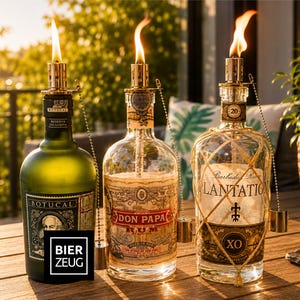 Könnte beinhalten: Drei Glasflaschen von Rum, die als Ölfackeln mit brennenden Dochten umfunktioniert wurden. Die Flaschen tragen Etiketten von Botucal, Don Papa und Plantation XO. Jede Flasche hat einen goldfarbenen Metallfackelaufsatz mit Kette und Gewicht.