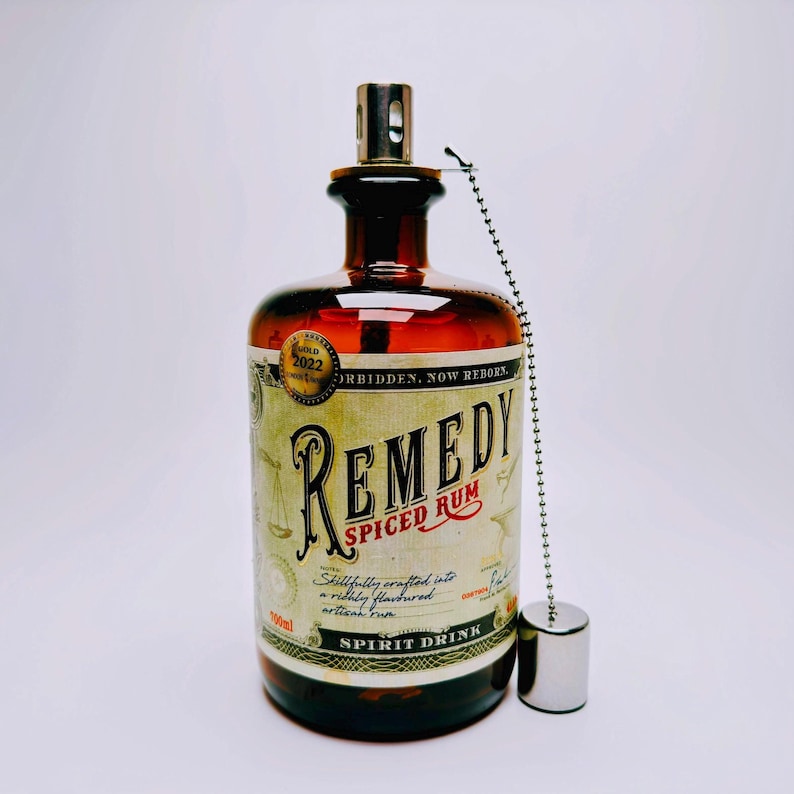 May include: A brown glass bottle of Remedy Spiced Rum with a metal wick assembly and a silver chain attached to a cap. The label features the text Remedy Spiced Rum, 700ml, and a gold 2022 award seal. The bottle has a wide base and a narrow neck.