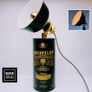 Może przedstawiać: Czarna i złota lampa biurkowa wykonana z puszki po whisky Aberfeldy 15 Highland Single Malt Scotch. Lampa posiada czarny klosz z białym wnętrzem, złote regulowane ramię oraz czarno-biały sznur. Tekst "BIER ZEUG".