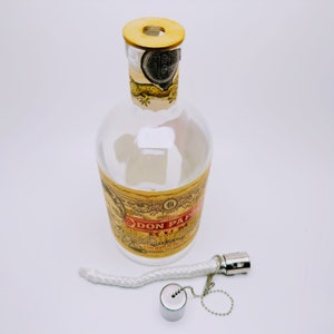 May include: A clear glass bottle with a gold label that reads "Don Papa Rum" and a gold cap. The bottle is sitting on a white surface. A white wick and a silver metal cap with a chain are also visible.
