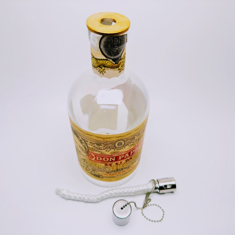 May include: A clear glass bottle with a gold label that reads "Don Papa Rum" and a gold cap. The bottle is sitting on a white surface. A white wick and a silver metal cap with a chain are also visible.
