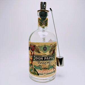May include: A clear glass bottle of Don Papa Baroko rum with a beige label featuring intricate illustrations of a moth, a figure, and foliage. The bottle has a metallic cap attached to a silver ball chain that hangs down the side of the glass.