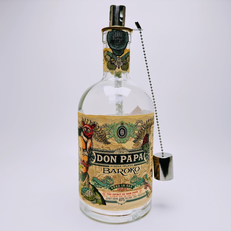 May include: A clear glass bottle of Don Papa Baroko rum with a beige label featuring intricate illustrations of a moth, a figure, and foliage. The bottle has a metallic cap attached to a silver ball chain that hangs down the side of the glass.